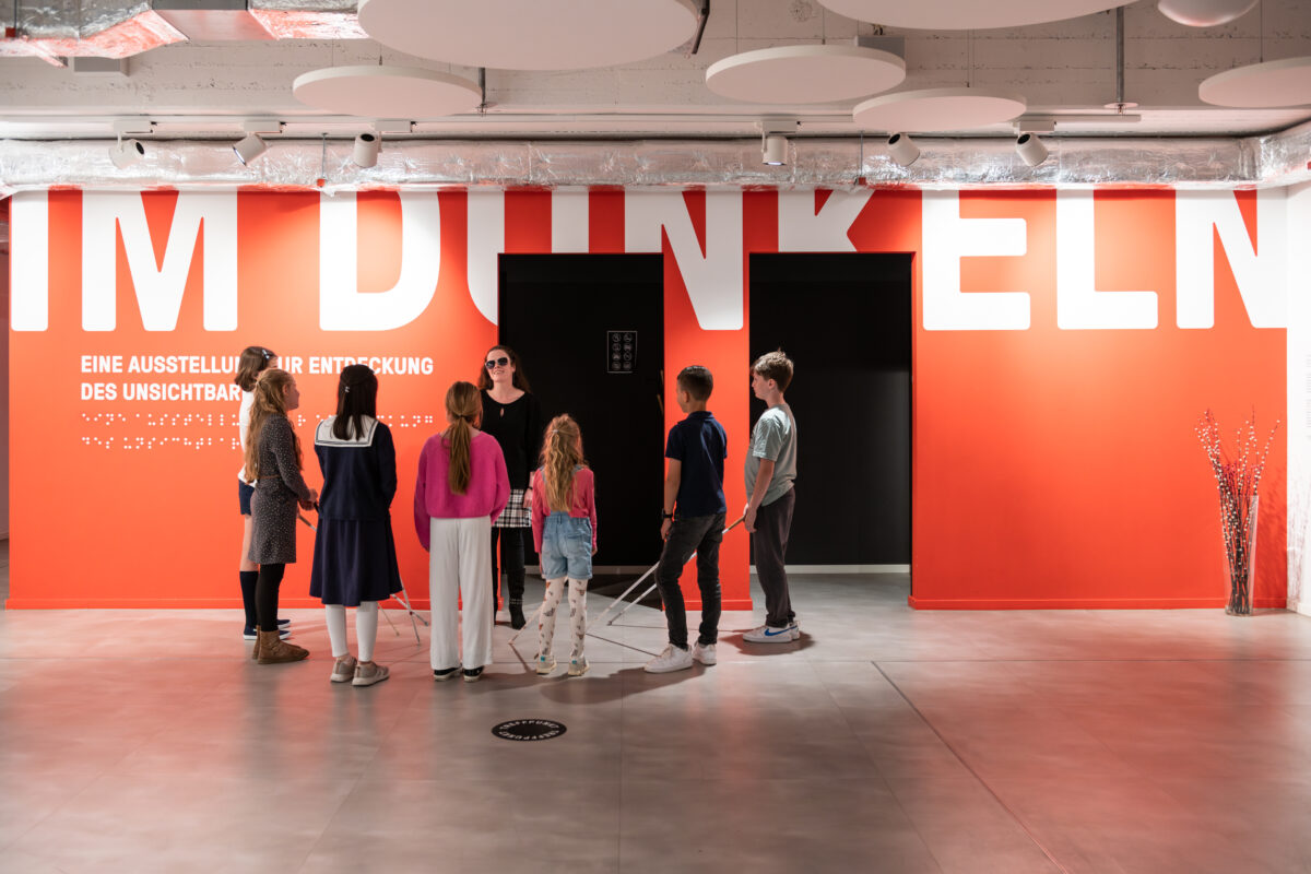 View into the foyer with a red wall with the inscription: "IM DUNKELN" in white letters. In front of it is a group of children standing around a blind woman.