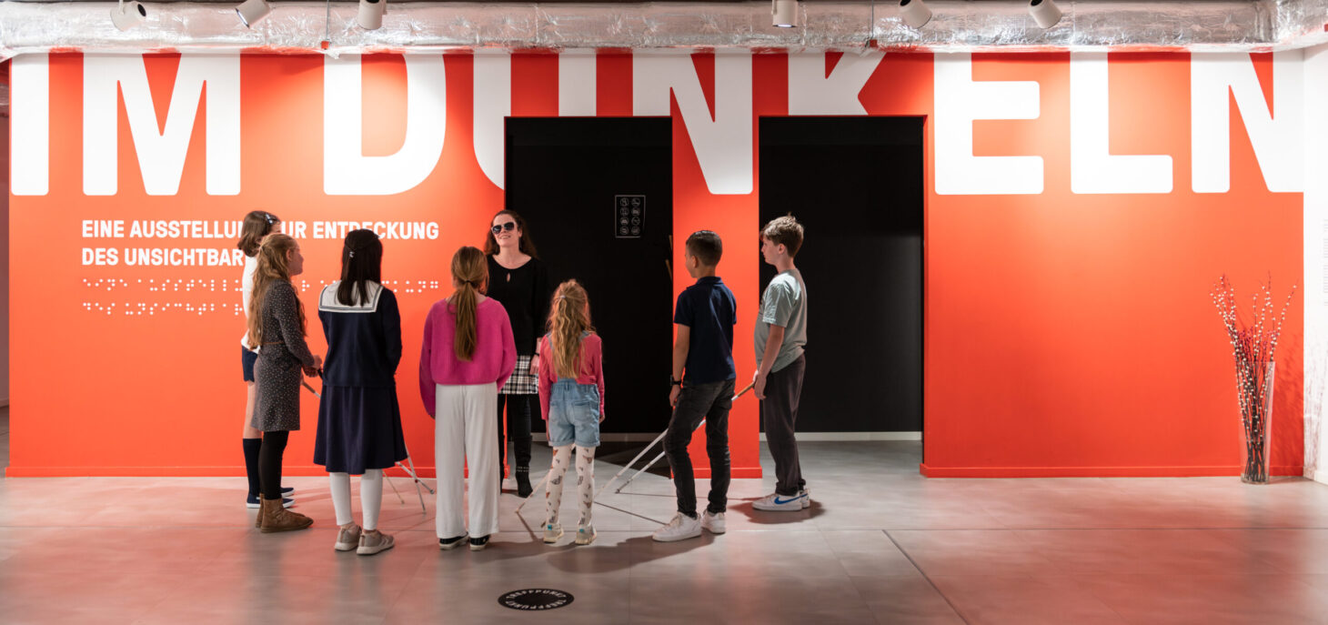 View into the foyer with a red wall with the inscription: "IM DUNKELN" in white letters. In front of it is a group of children standing around a blind woman.