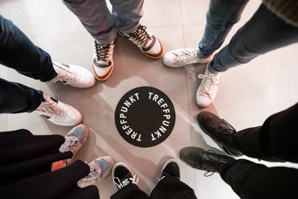 Photo from above of six pairs of feet standing in a circle around a black dot on the floor. On the black dot is written in white letters: "Treffpunkt". ("Meeting point")