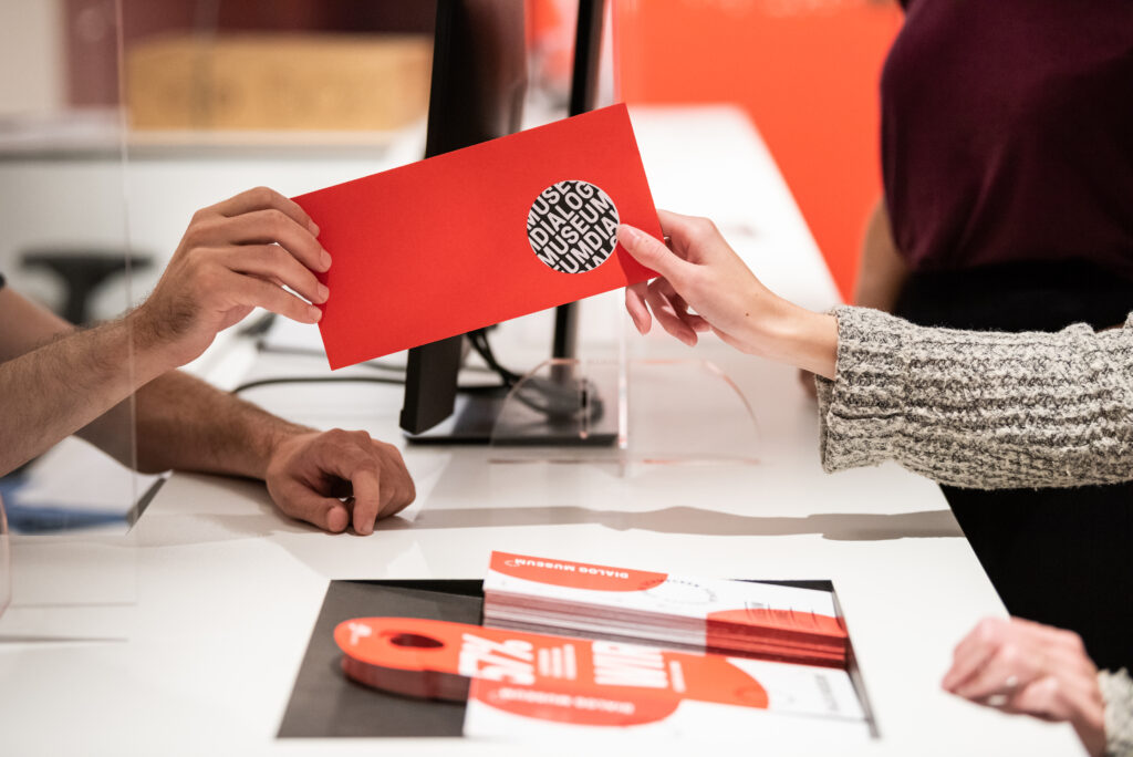 Above the museum counter, a hand passes a red envelope with a round Dialogue Museum logo to another person.