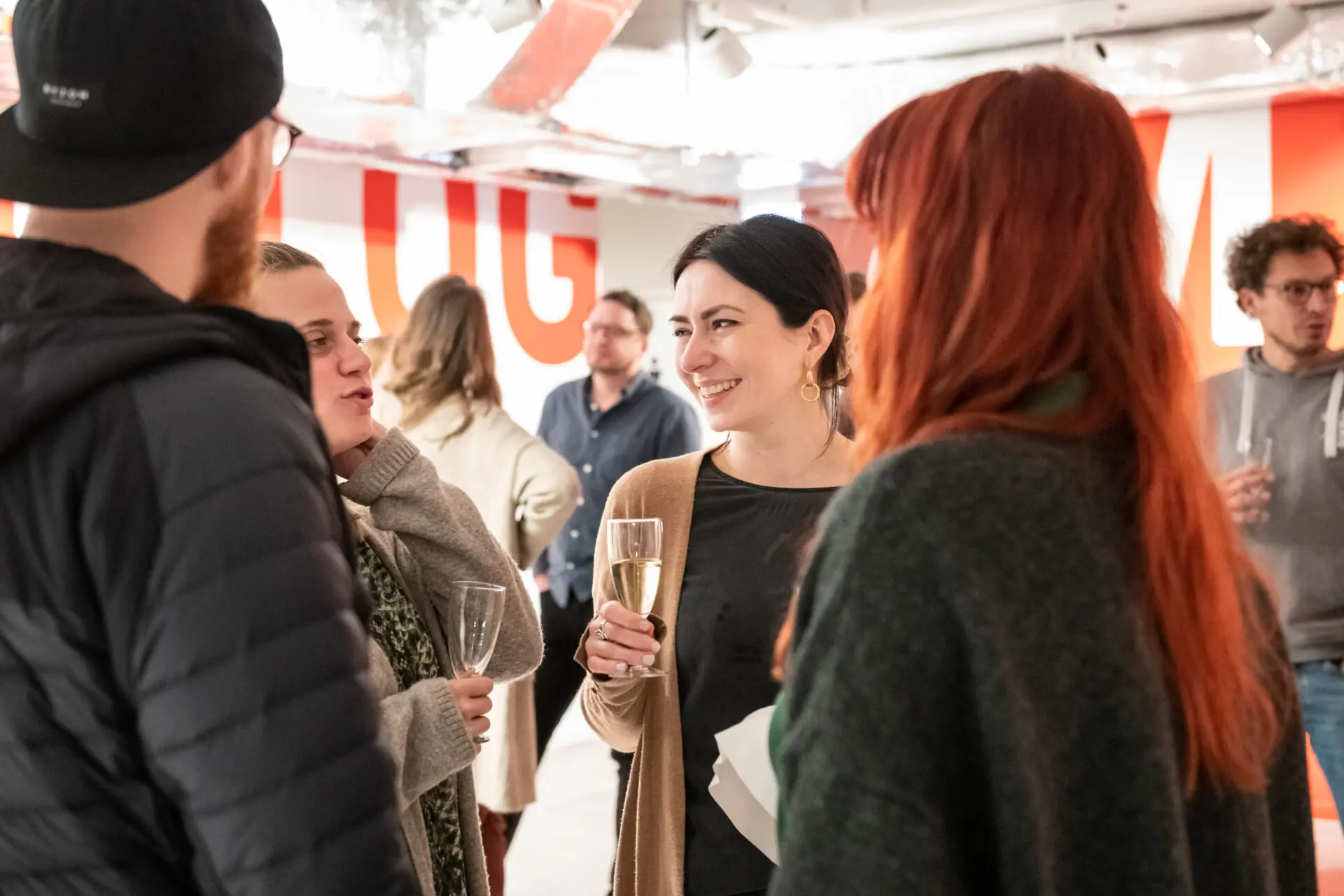 You can see the illuminated foyer of the Dialogue Museum. In the background, several people are standing in small groups, engaged in conversation. In the foreground of the picture, four people are deep in conversation. The focus is on a laughing woman with dark hair tied back. Two people are holding filled champagne glasses in their hands.