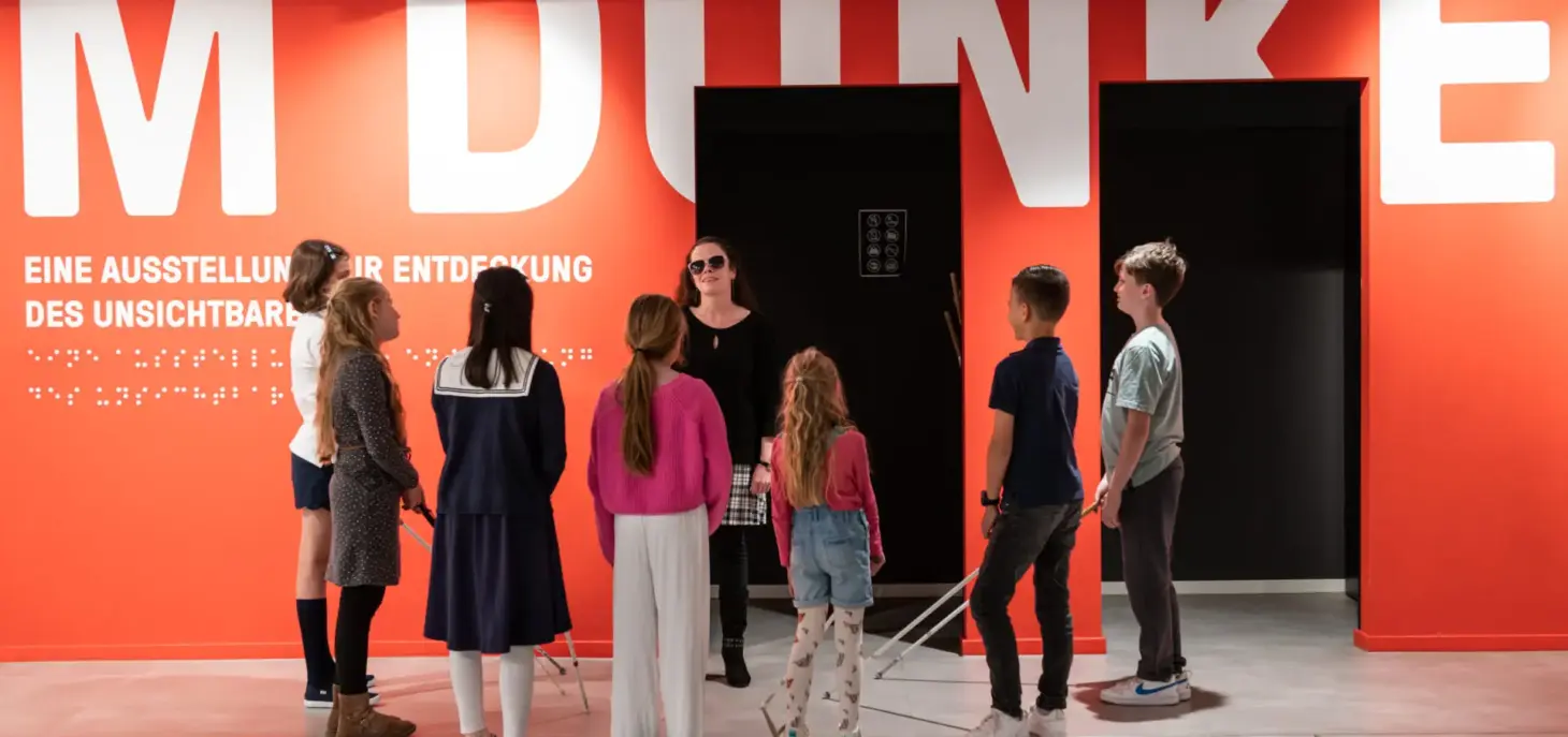 View into the foyer with a red wall with the inscription: "IM DUNKELN" in white letters. In front of it is a group of children standing around a blind woman.