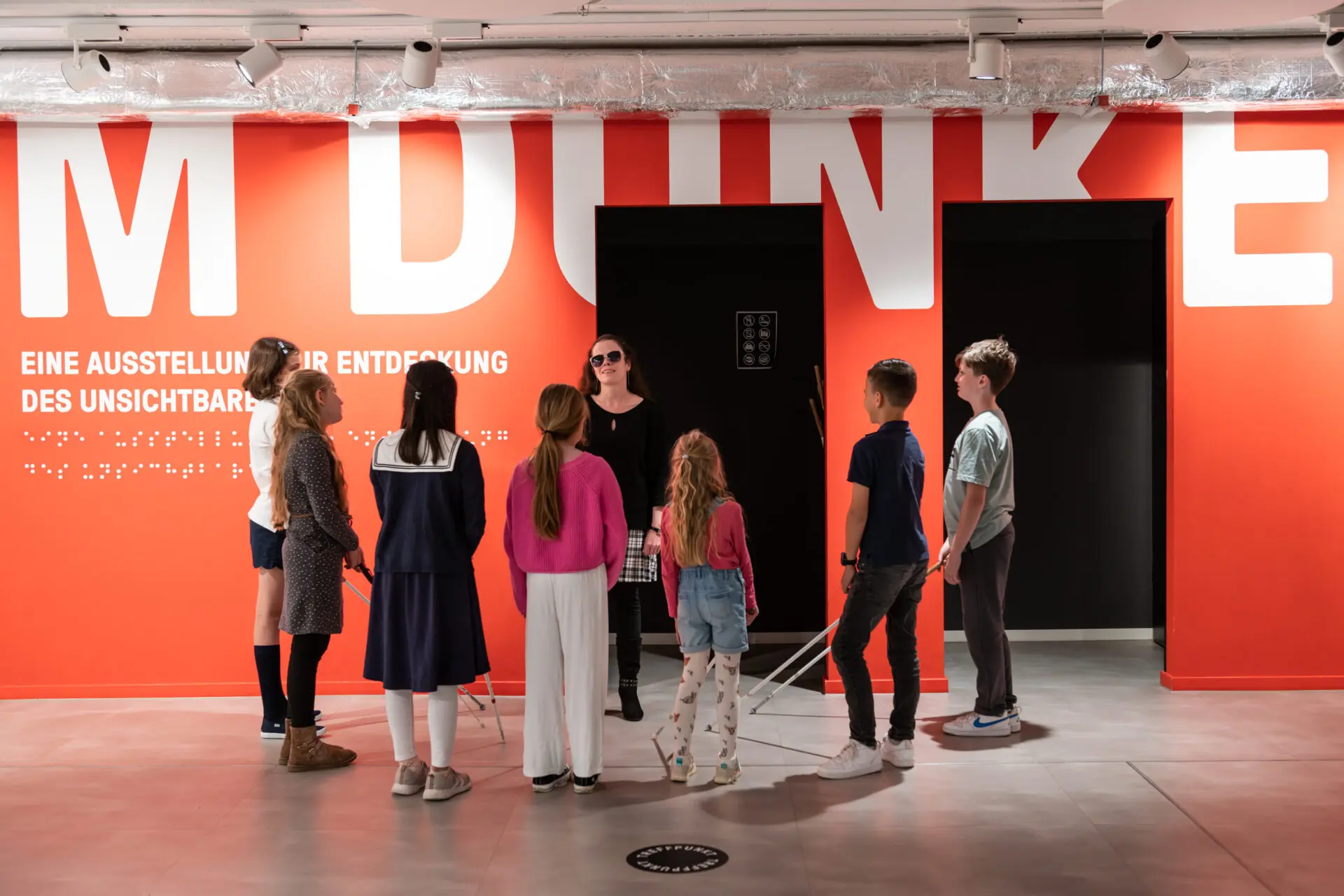 View into the foyer with a red wall with the inscription: "IM DUNKELN" in white letters. In front of it is a group of children standing around a blind woman.