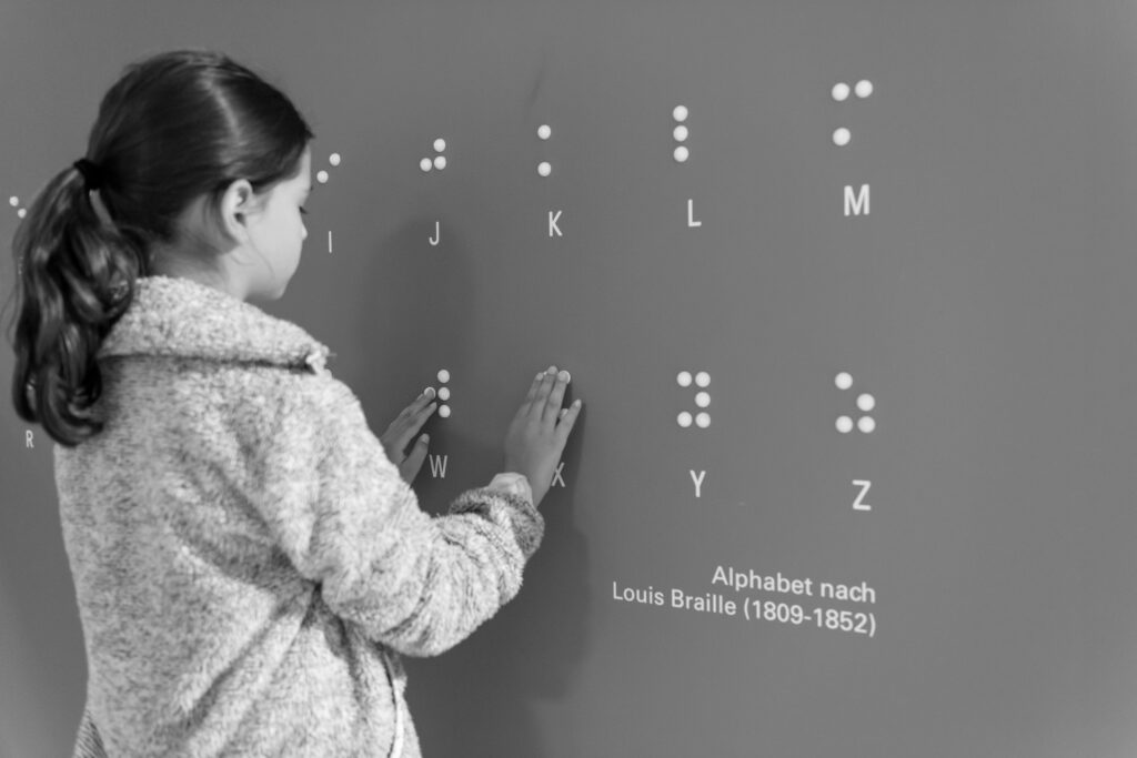 Ein Mädchen tastet über ein fühlbares Braille-Alphabet an der Museumswand.