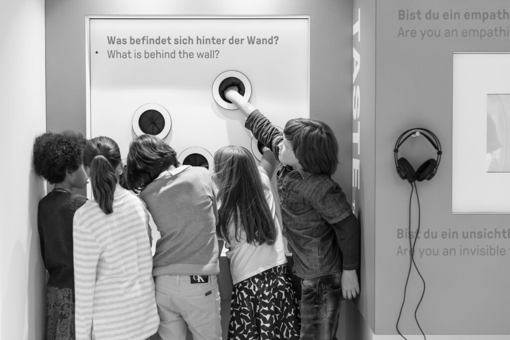 Kinder stehen vor einer Tast-Wand und stecken ihre Hände durch Löcher darin, um zu fühlen was dahinter ist.