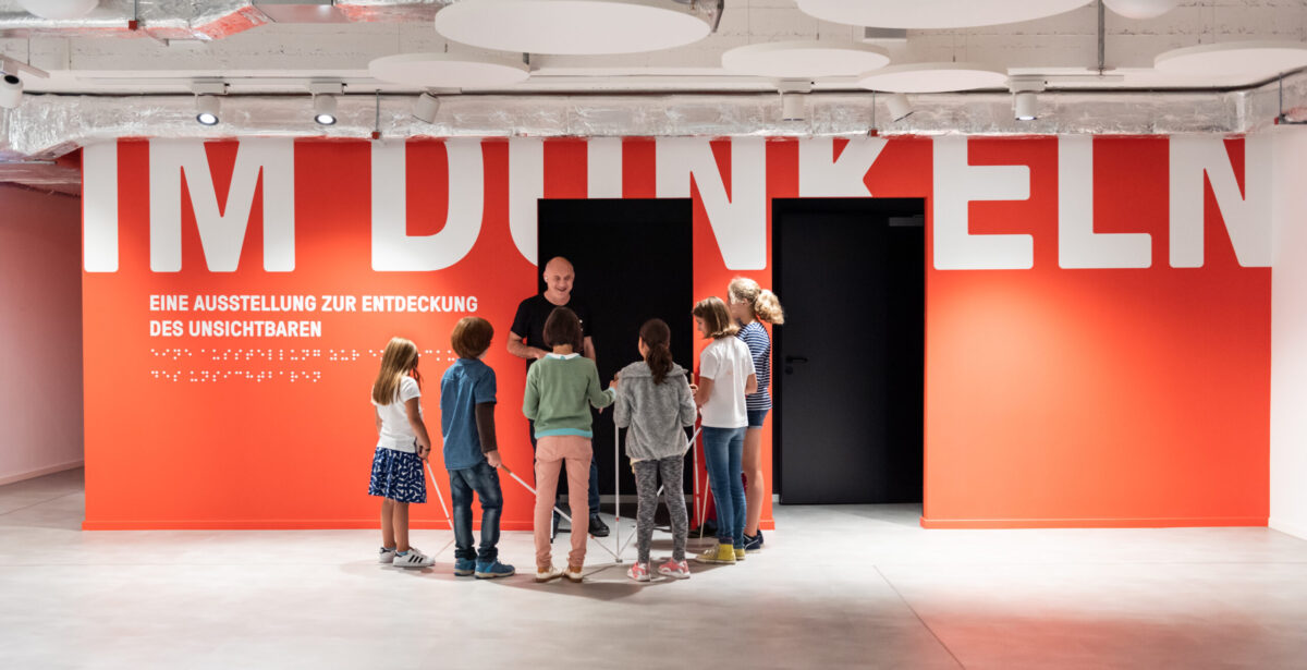 Blick in das Foyer mit einer roten Wand mit der Aufschrift: "IM DUNKELN" in weißen Letter. Davor steht eine Gruppe von Kindern die um einen blinden Mann stehen.
