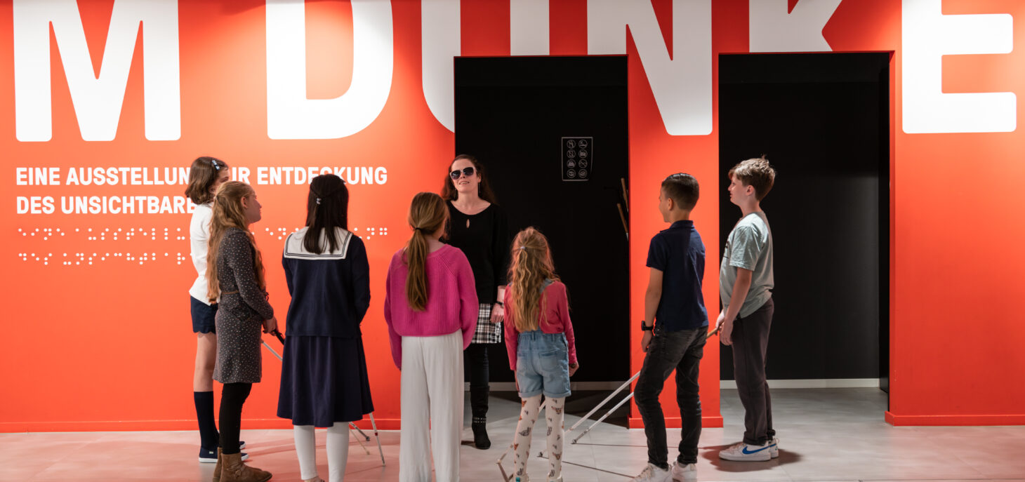 Blick in das Foyer mit einer roten Wand mit der Aufschrift: "IM DUNKELN" in weißen Letter. Davor steht eine Gruppe von Kindern die um eine blinde Frau stehen.