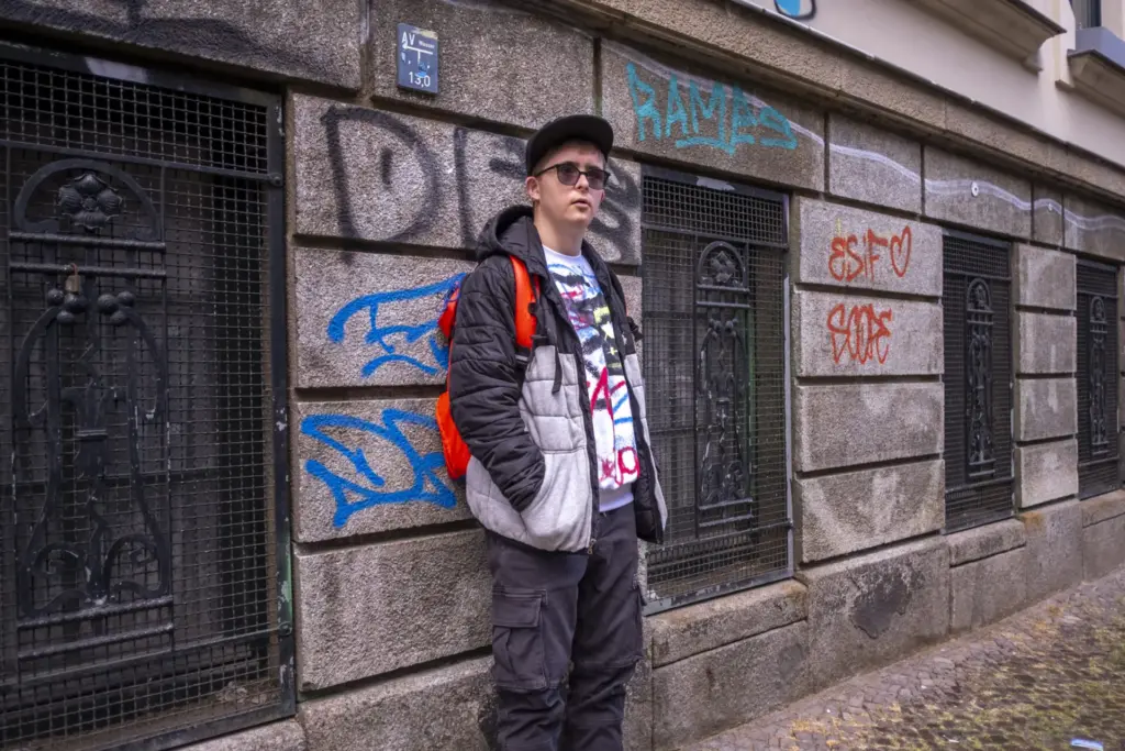 Ein Junge steht vor einer Hauswand. Er trägt eine Sonnenbrille, schwarze Cappi, Daunenjacke und einen leuchtend orangenen Rucksack.