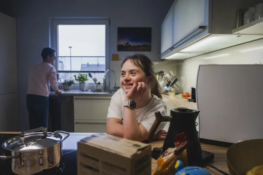 Ein Mädchen lehnt sich auf eine Küchenzeile, um sie herum stehen Küchenutensilien. Sie schaut aus dem Bild raus und lächelt. Im Hintergrund steht eine Frau mit kurzen Haaren an der Spülmaschine.